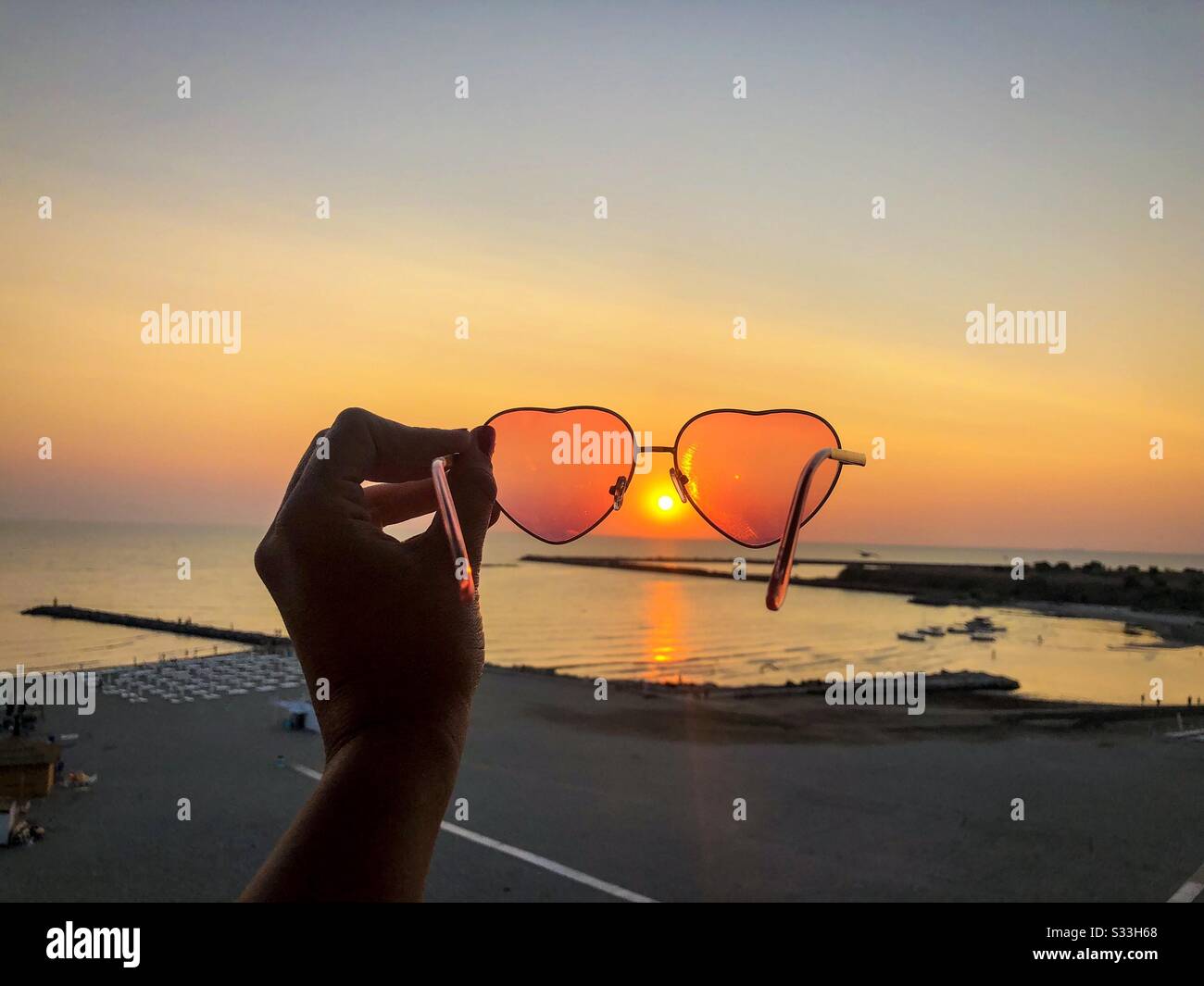 Woman holding heart shape sunglasses over the beach at sunset - Smartphone Captured Stock Image