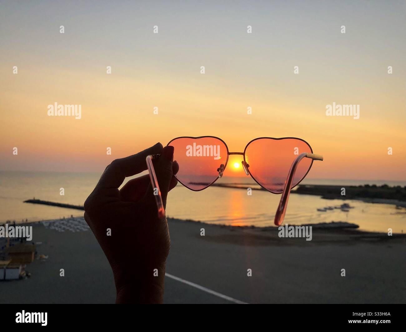 Woman holding heart shape sunglasses over the beach at sunset - Smartphone Captured Stock Image