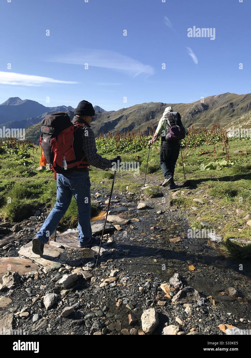 Two men hiking through a small stream bed with backpacks on in a meadow surrounded by mountains and a blue sky. - Smartphone Captured Stock Image Two men hiking through a small stream bed with backpacks on in a meadow surrounded by mountains and a blue sky. - Smartphone Captured Stock Image