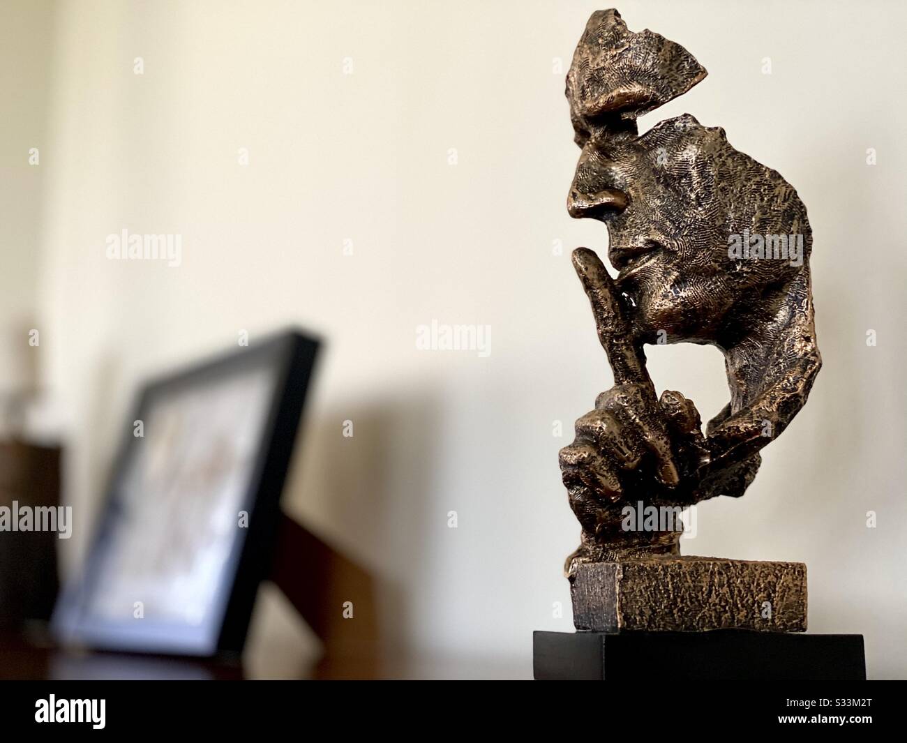 A close up high contrast shot of a modern head statue on a home sideboard with blurry picture frame in background. Finger gesture for peace and quiet. Finger raised to mouth. Shh - Smartphone Captured Stock Image