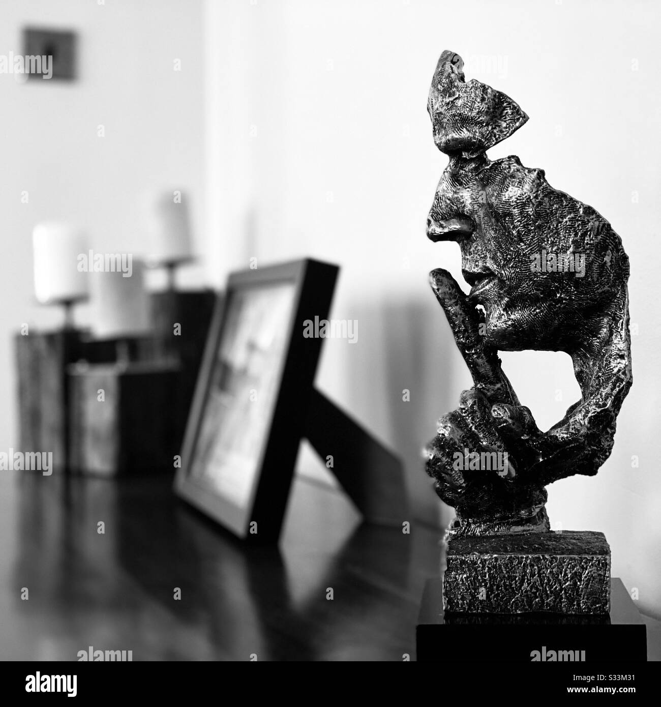 A close up monochrome view of a modern home ornament on a wooden sideboard. Abstract male head ornament with finger raised to mouth. Quiet in living room - Smartphone Captured Stock Image