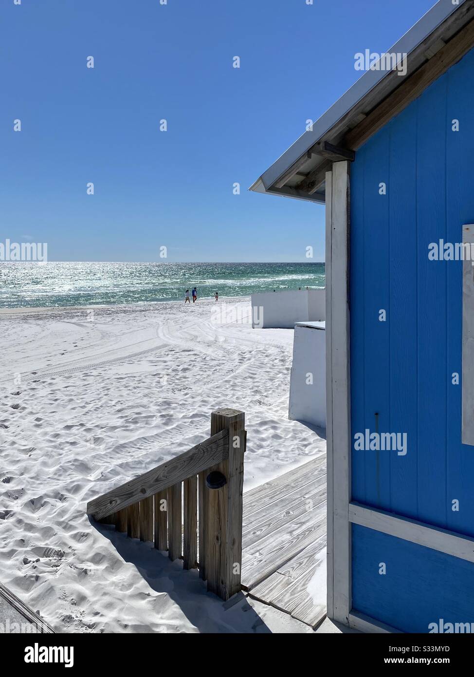 Colorful blue beach hut entrance to white sand beach with sun sparkling on the emerald ocean water - Smartphone Captured Stock Image