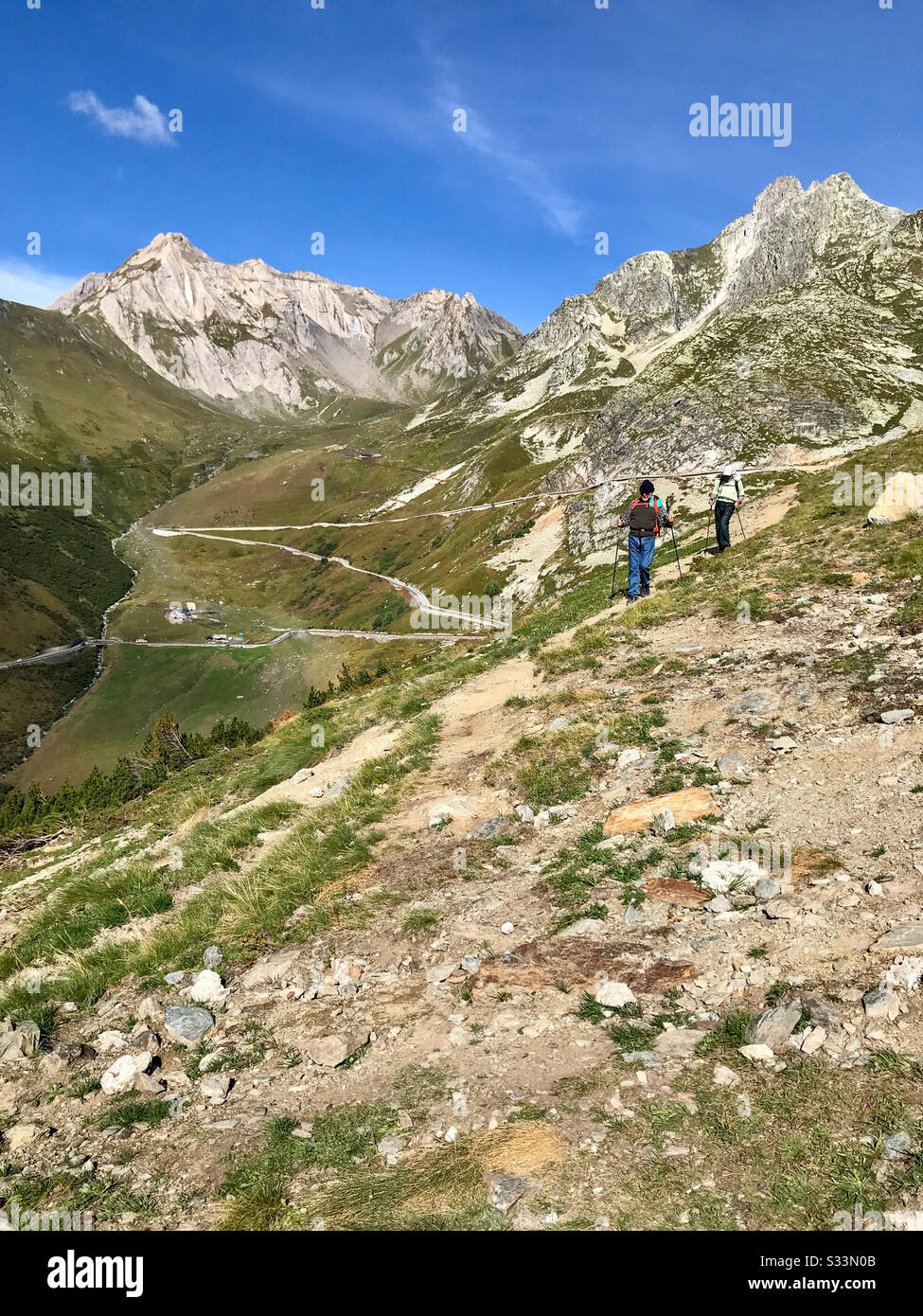 Two men with backpacks and walking sticks hiking the Via Francigena steep trail in the Italian Alps. Beautiful blue sky, rocky zigzag terrain. - Smartphone Captured Stock Image