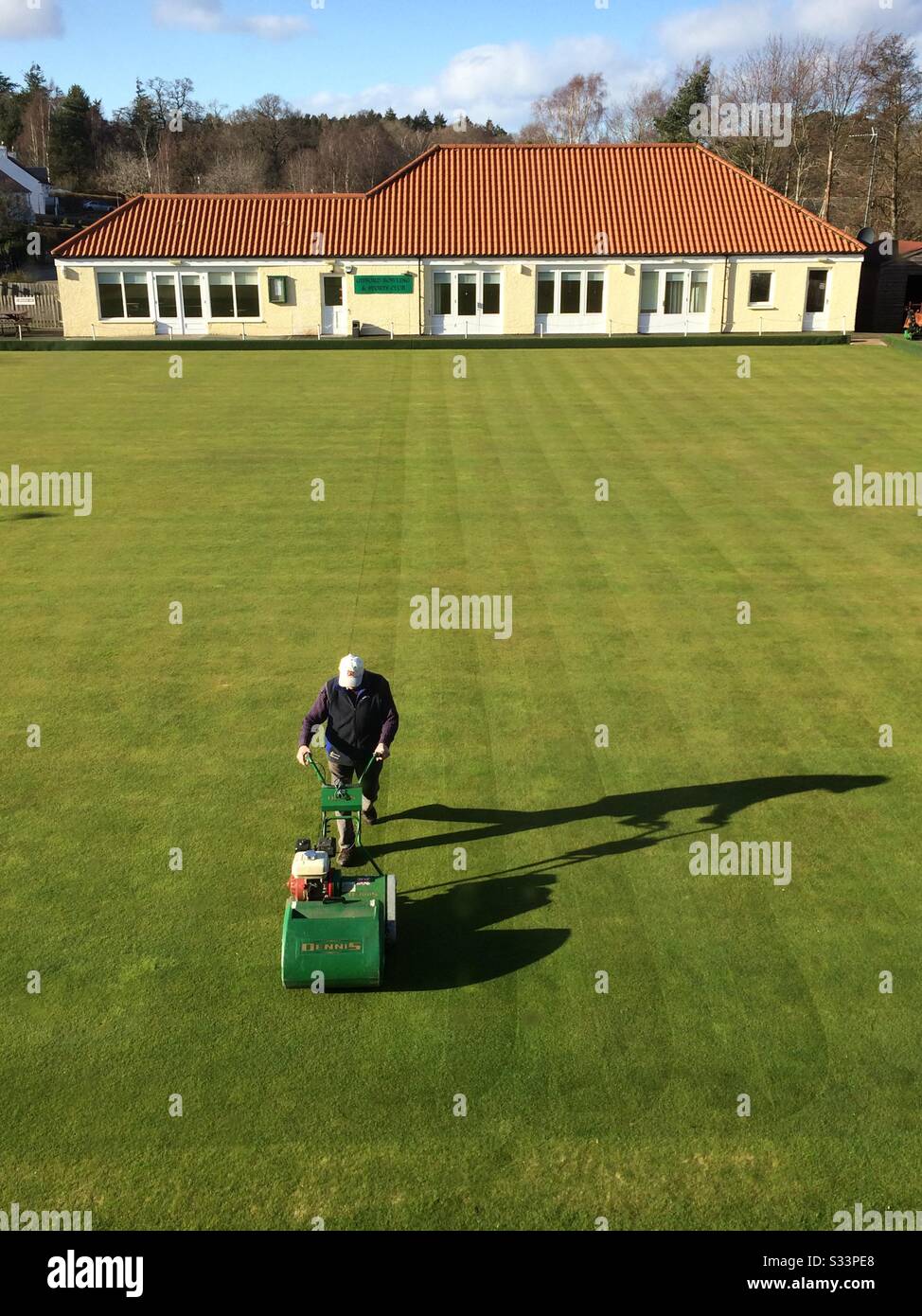 The first cut of spring at the Bowling Club in Gifford, East Lothian, UK. With dusk approaching, after a dry day, a volunteer official is out mowing with only his shadow for company. Stock Photo