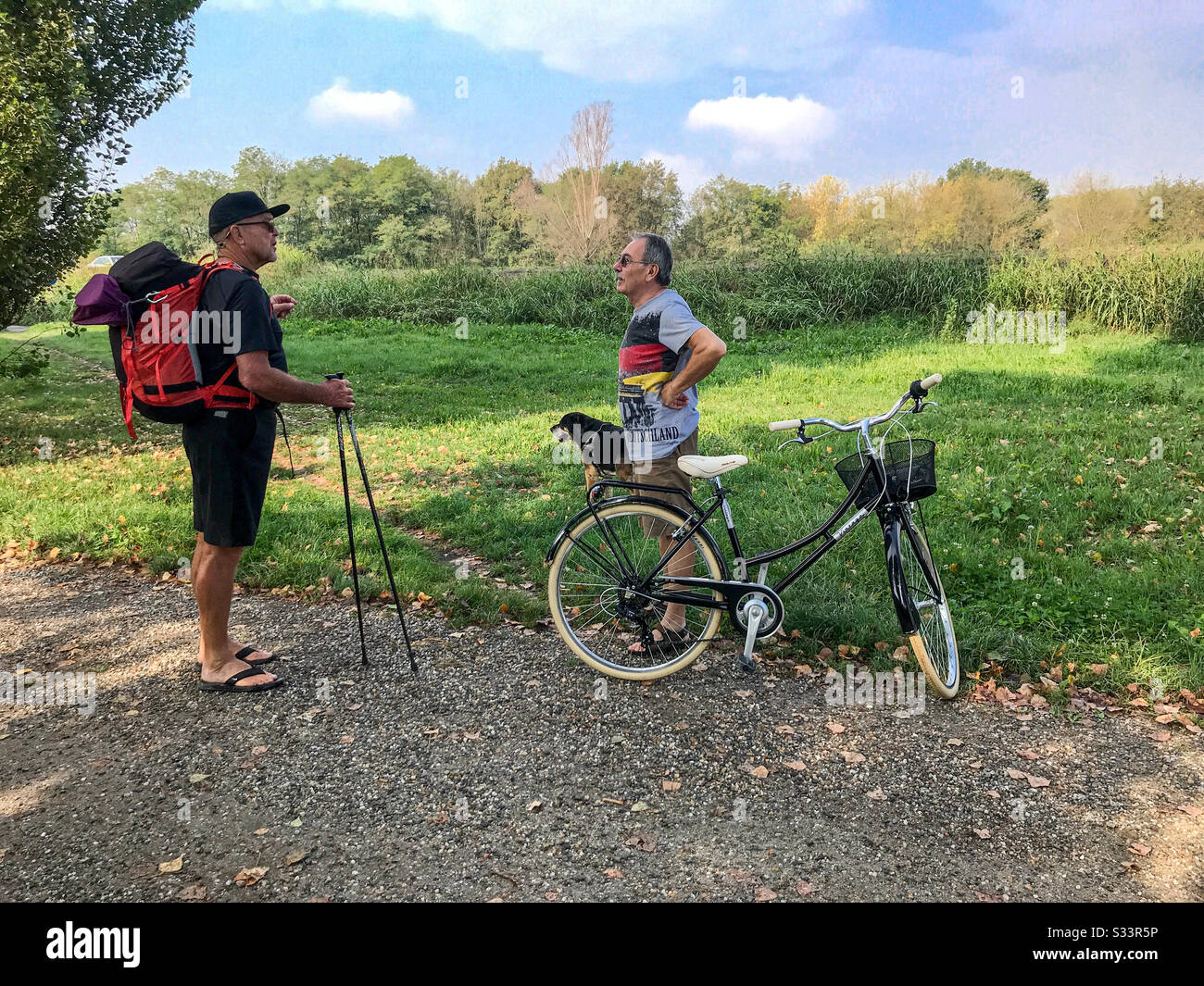 A countryside scene of a male backpacker talking to a local man standing next to a bicycle with his dog. - Smartphone Captured Stock Image