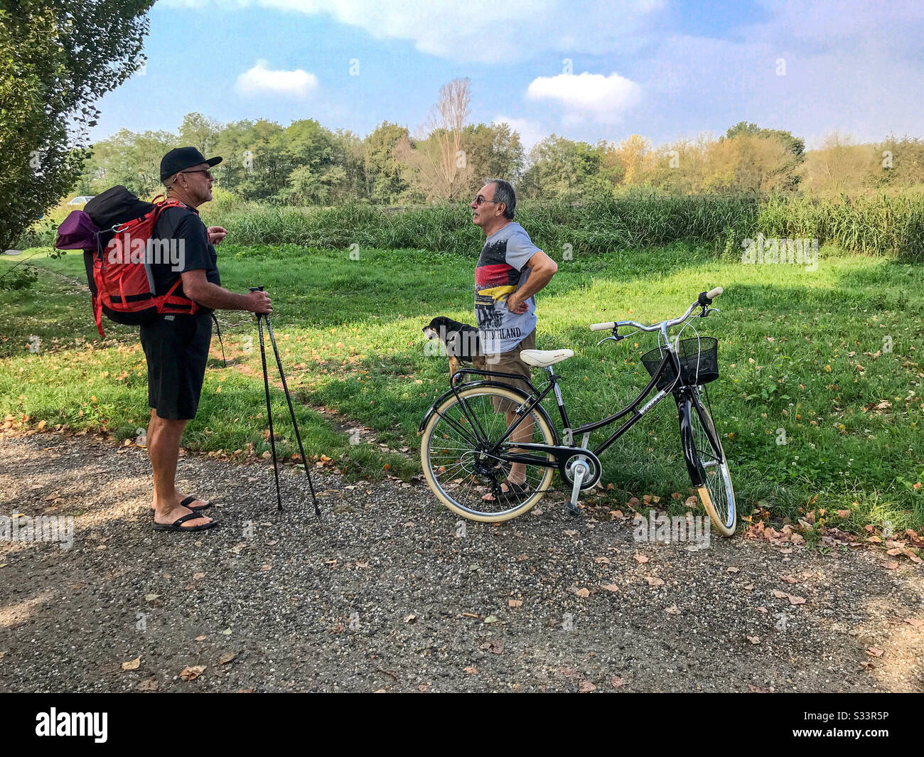 A countryside scene of a male backpacker talking to a local man standing next to a bicycle with his dog. Stock Photo