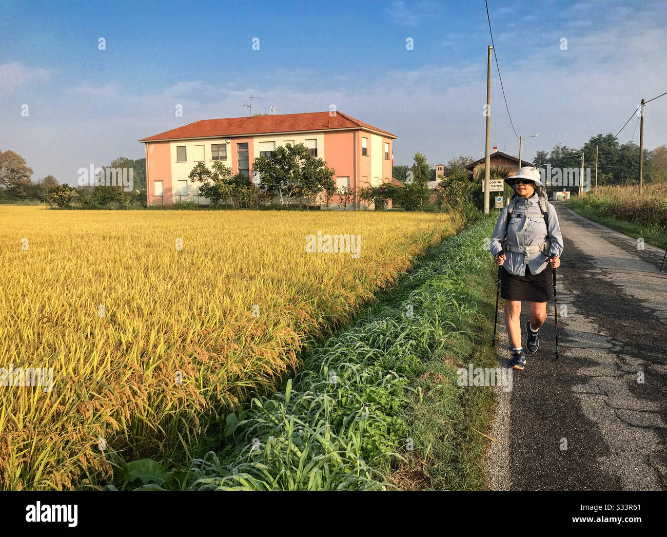 Female backpacker walking on a road next to a field of rice with an old vintage peach colored Italian house in the background. - Smartphone Captured Stock Image