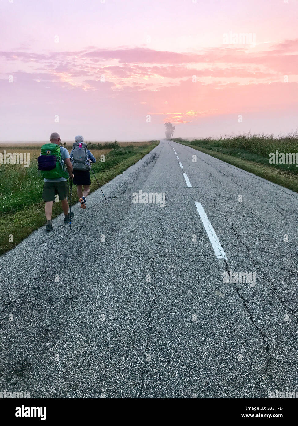 Husband and wife backpackers walking on a rural paved road surrounded by fields during sunrise. - Smartphone Captured Stock Image