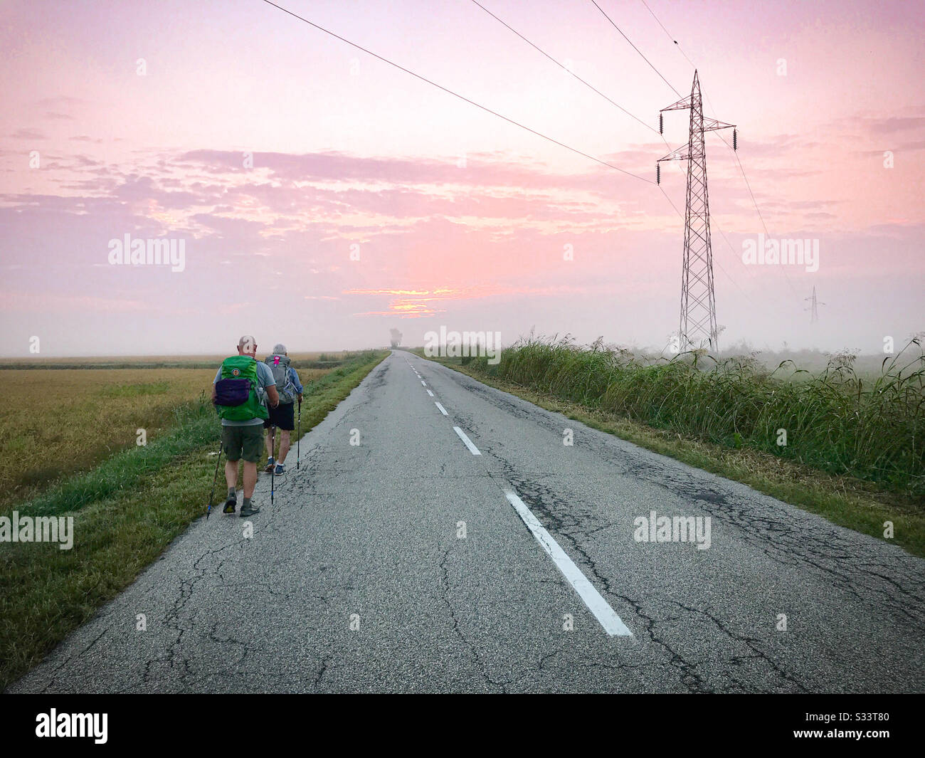 Two backpackers walking next to a rural paved road during sunrise next to a harvested rice field. A tall electrical tower is on the right side of the road. - Smartphone Captured Stock Image