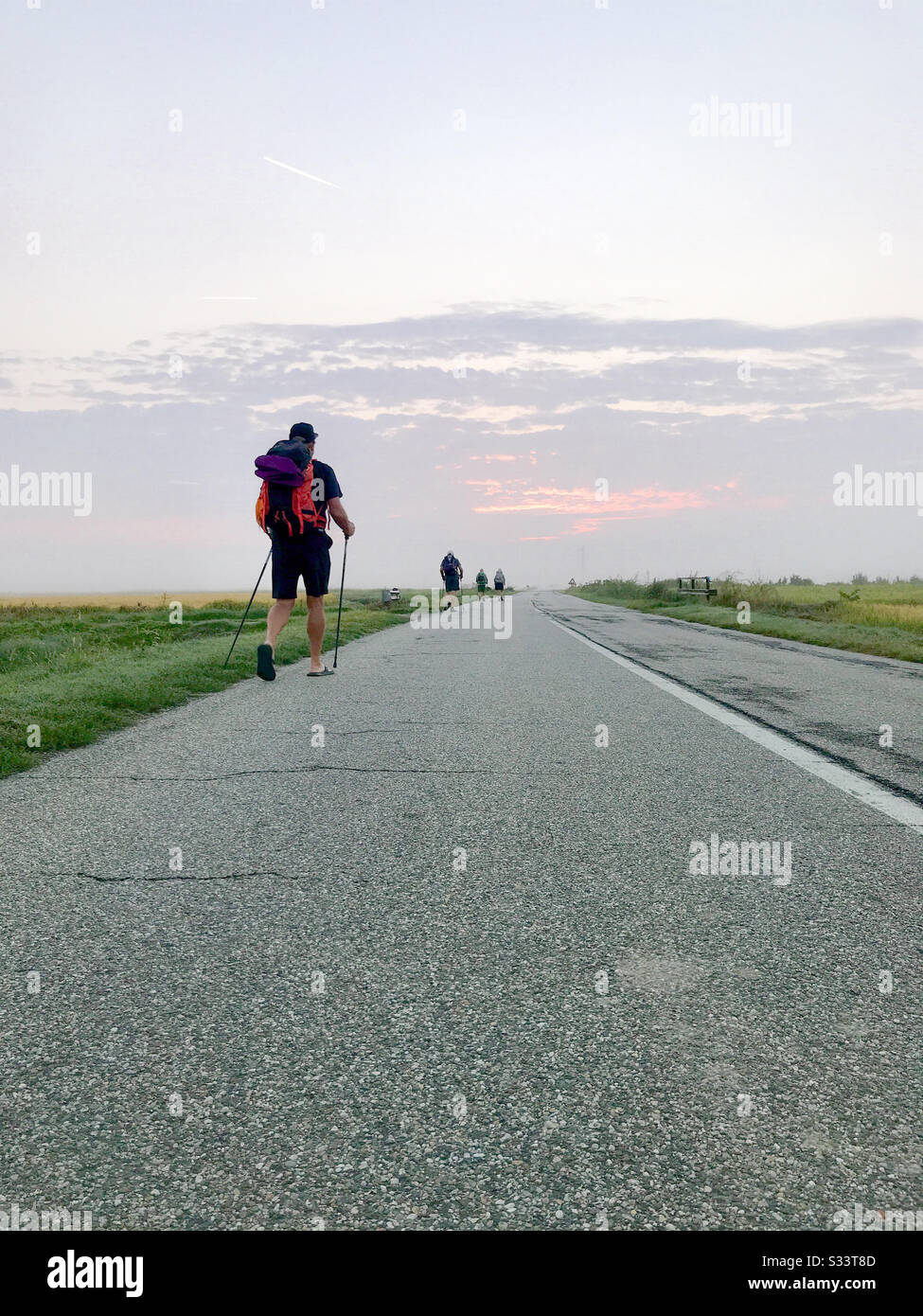 Backpackers walking next to a rural paved street taken from a lower angle. Cloudy sunrise. - Smartphone Captured Stock Image