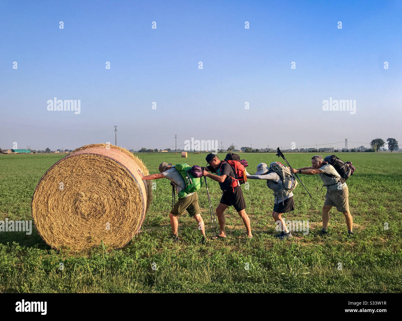 Four backpackers being silly by lining up and pushing on each other’s backs while trying to move a huge hay roll on an open field. - Smartphone Captured Stock Image