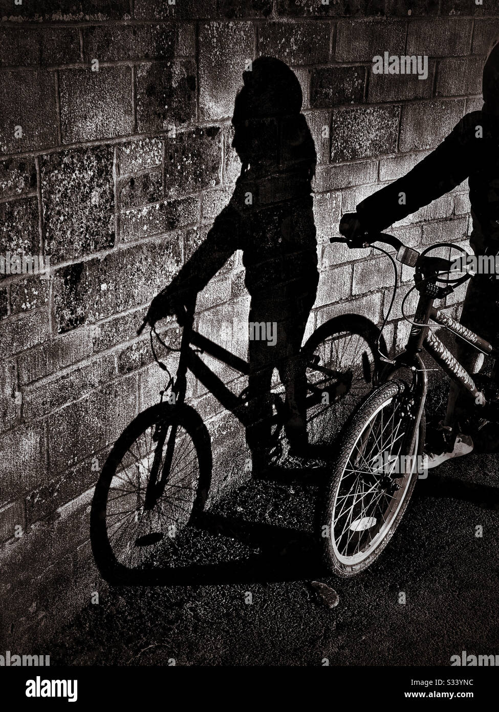 The shadow of a child riding a bike falls onto a tall block wall. An atmospheric picture that has multiple potential uses. Photo Credit - © COLIN HOSKINS. - Smartphone Captured Stock Image