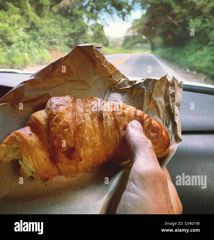 A fresh, flaky croissant is enjoyed inside a car on a road trip. Stock Photo