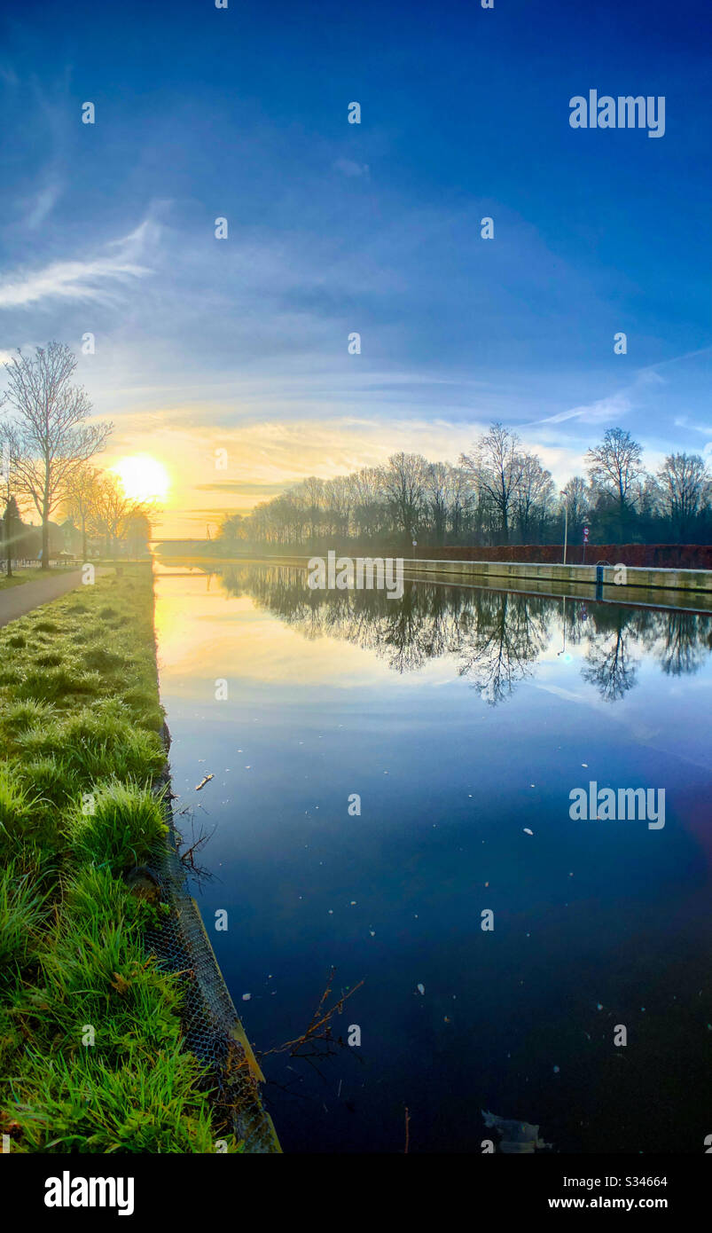 Beautiful golden sunrise in a clear deep blue sky over a forest reflected in the water of a canal or river, as seen from the grassy waterside - Smartphone Captured Stock Image