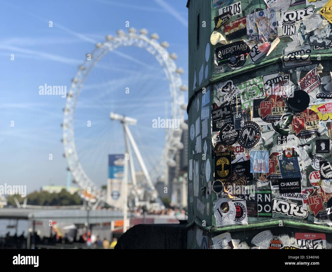 A selective focus shot of a lamppost covered in stickers at Westminster Pier with a blurry London Eye in the background - Smartphone Captured Stock Image