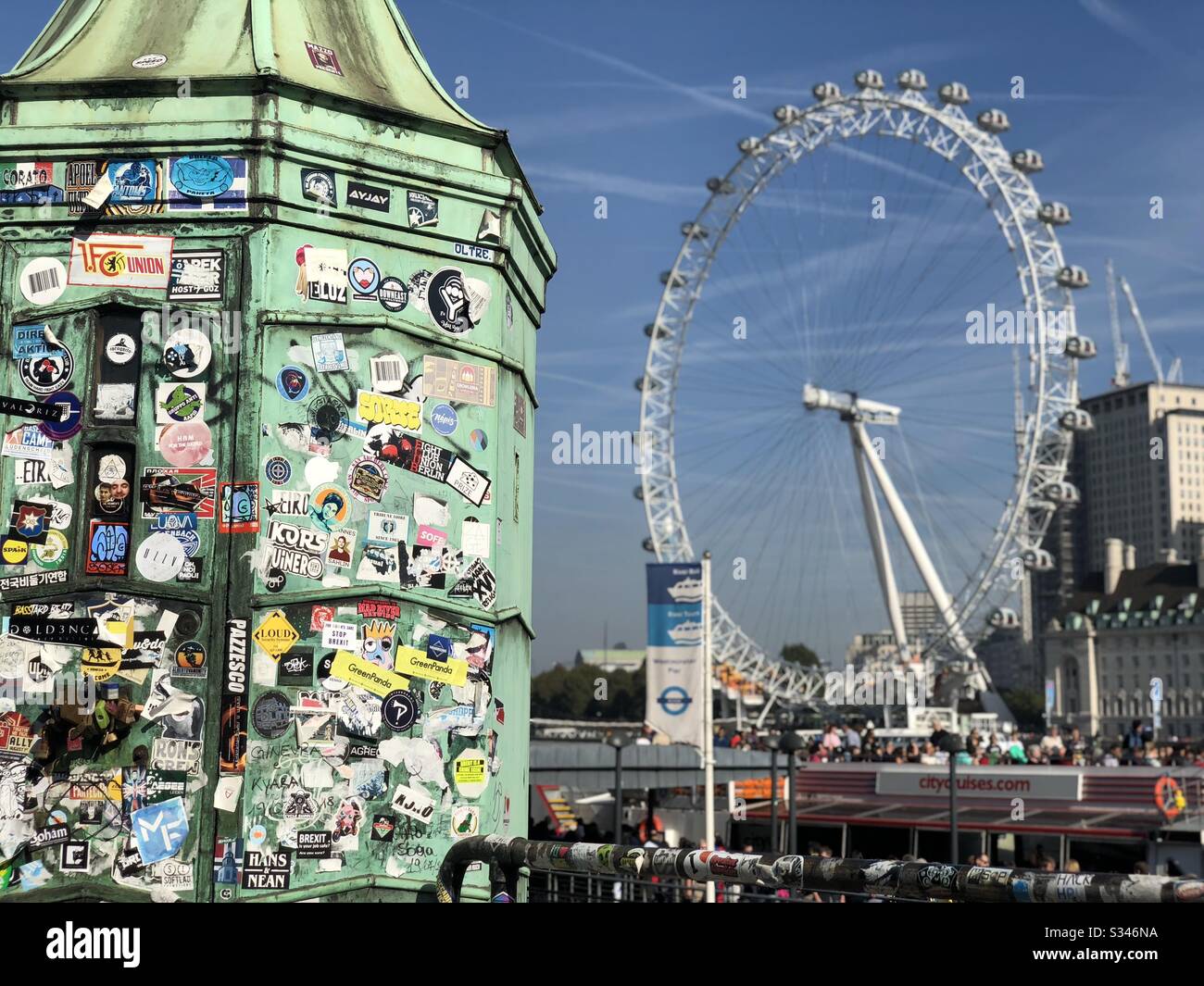 A close up shot of a lamppost covered in tourist stickers with a blurry Millennium Wheel in the background at Westminster Pier, London - Smartphone Captured Stock Image