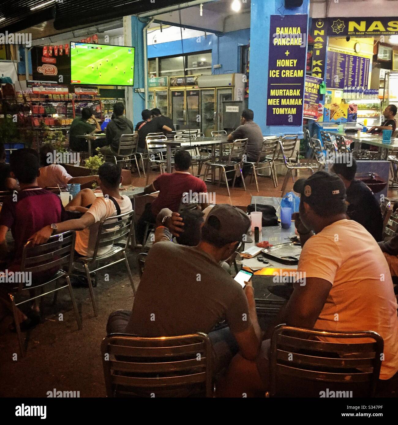 Local residents watch a soccer match on television outside an all-night restaurant in Batu Ferringhi, Penang, Malaysia - Smartphone Captured Stock Image