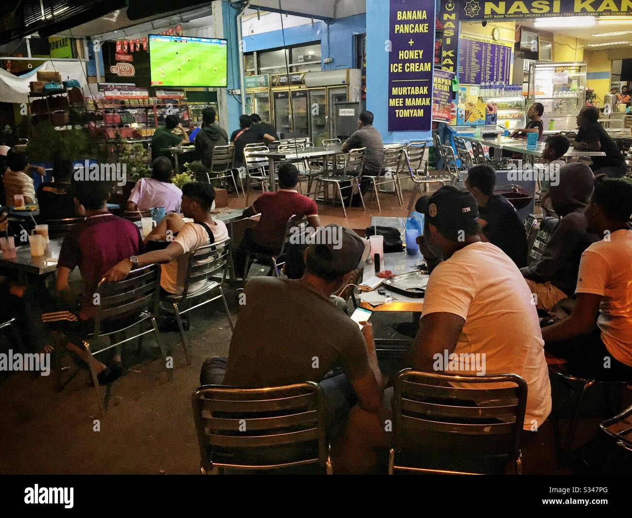 Local residents watch a soccer match on television outside an all-night restaurant in Batu Ferringhi, Penang, Malaysia - Smartphone Captured Stock Image