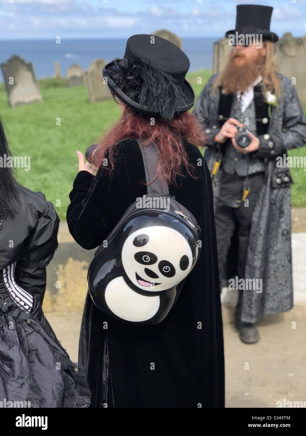 A lady is seen wearing goth clothes and a panda backpack from behind during a cosplay event on a graveyard. - Smartphone Captured Stock Image