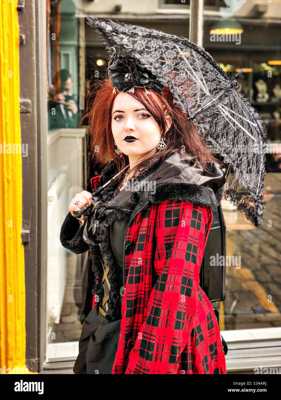 Person in a creative costume during a cosplay event. Whitby Goth Weekend. Girl with red and black gothic outfit, pale white face and black lace umbrella - Smartphone Captured Stock Image