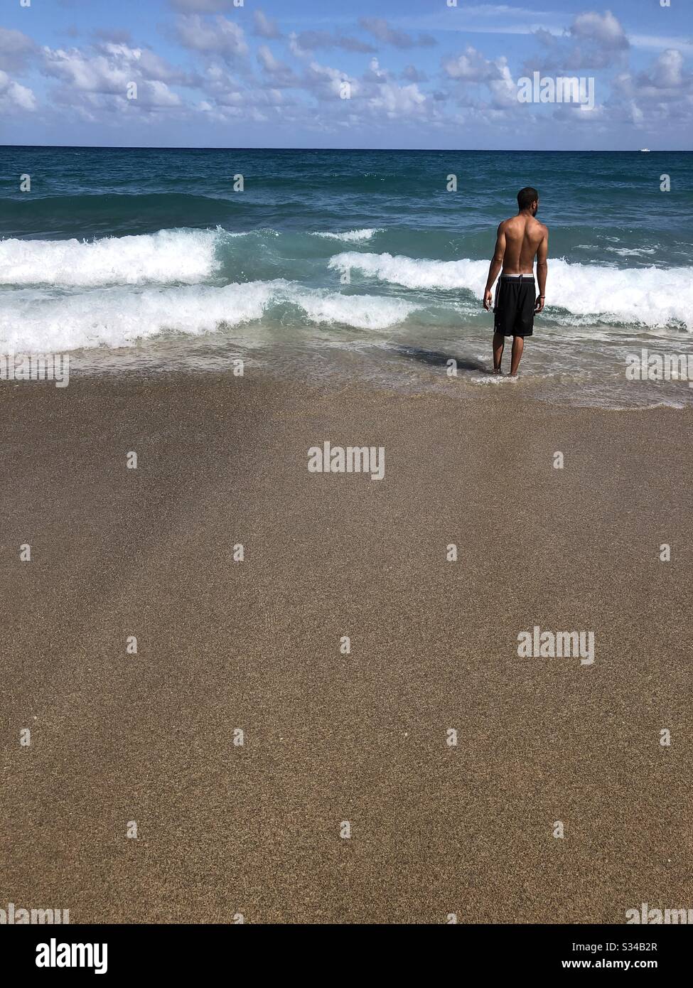 A young man standing on the sand looking out at the ocean in Lake Worth Beach, Florida. - Smartphone Captured Stock Image