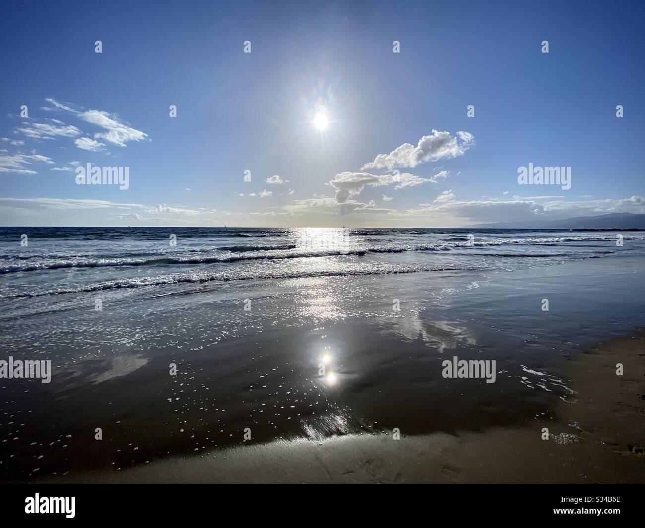 Centered landscape view of low sun over the Pacific Ocean with sparse clouds in blue sky, gentle waves lapping sandy beach in foreground - Smartphone Captured Stock Image