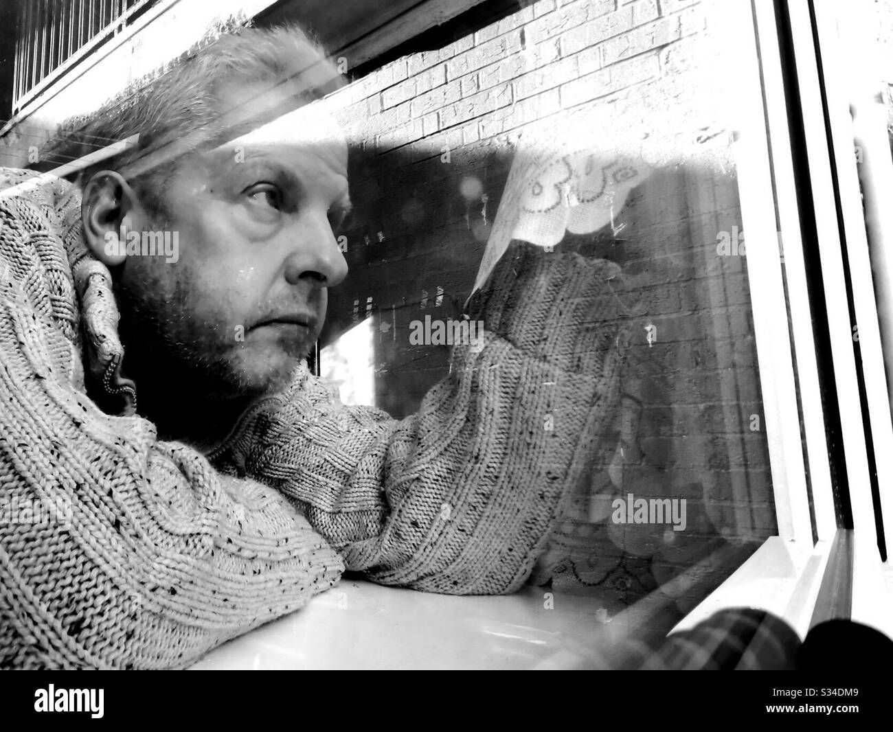Black and white photo of middle aged man pulling curtain aside and looking out of his window Stock Photo