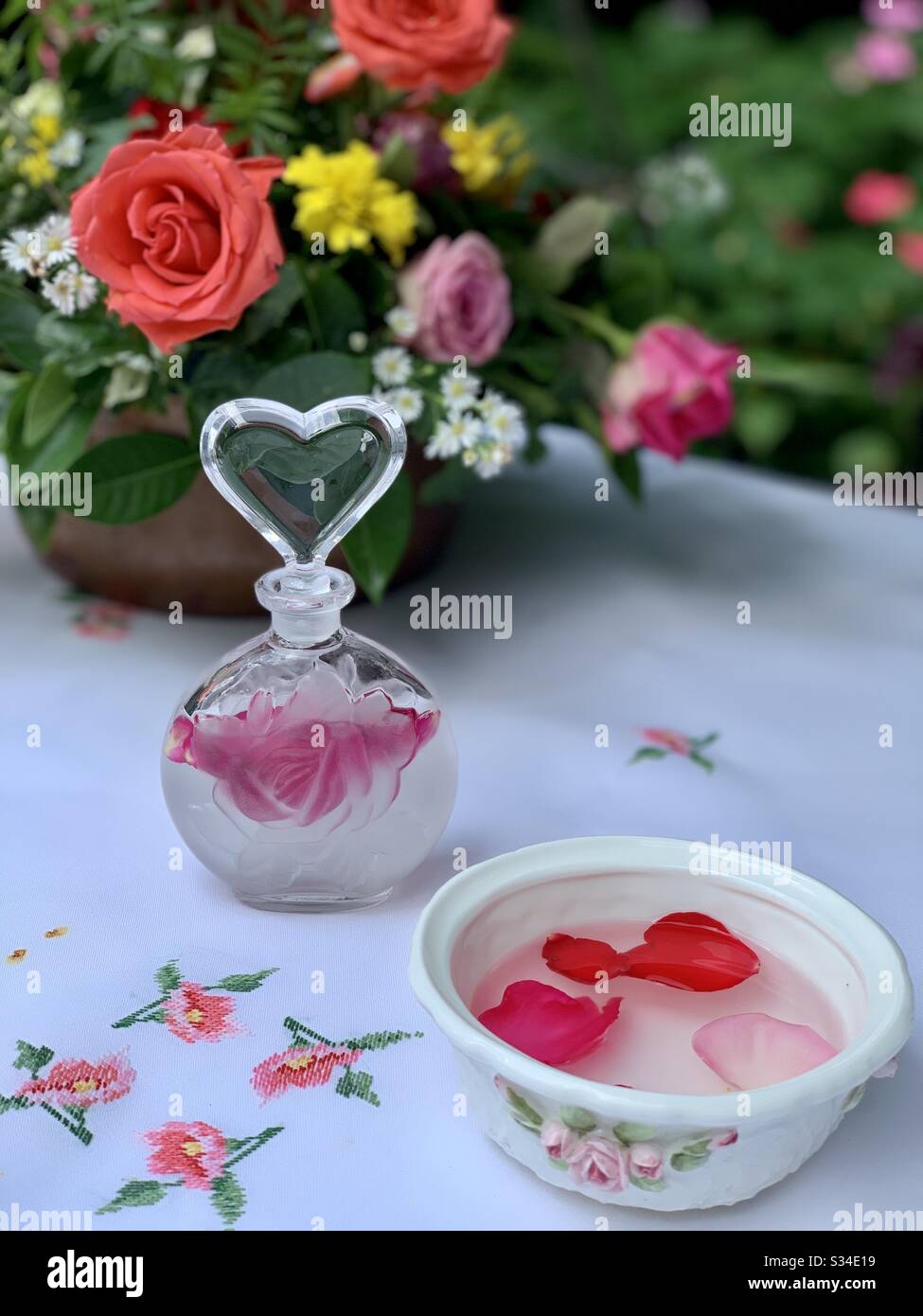 The delicate beauty of a charming perfume bottle filled with rose water. A bone China ramekin is filled with the rose petals and water that perfumes the air and the hands. Dainty linen and roses. Stock Photo