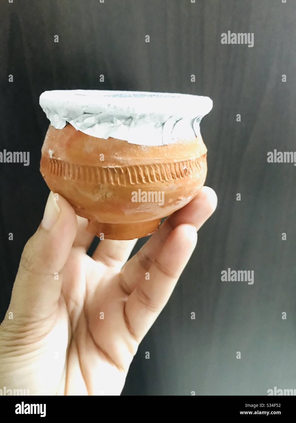 Hand holding designed Kulfi clay pot covered with aluminium foil against black rough surface-frozen dairy dessert originated from India, traditional Indian ice cream in earthen pot- Stock Photo