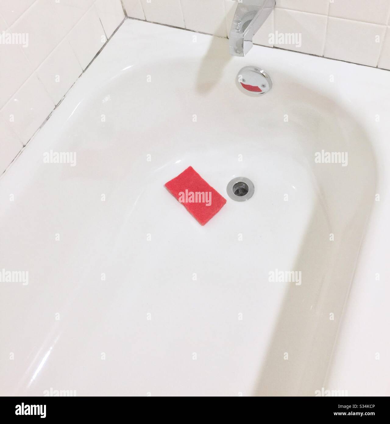 Gleaming white bath tub with red scour sponge near bath drain. Stock Photo