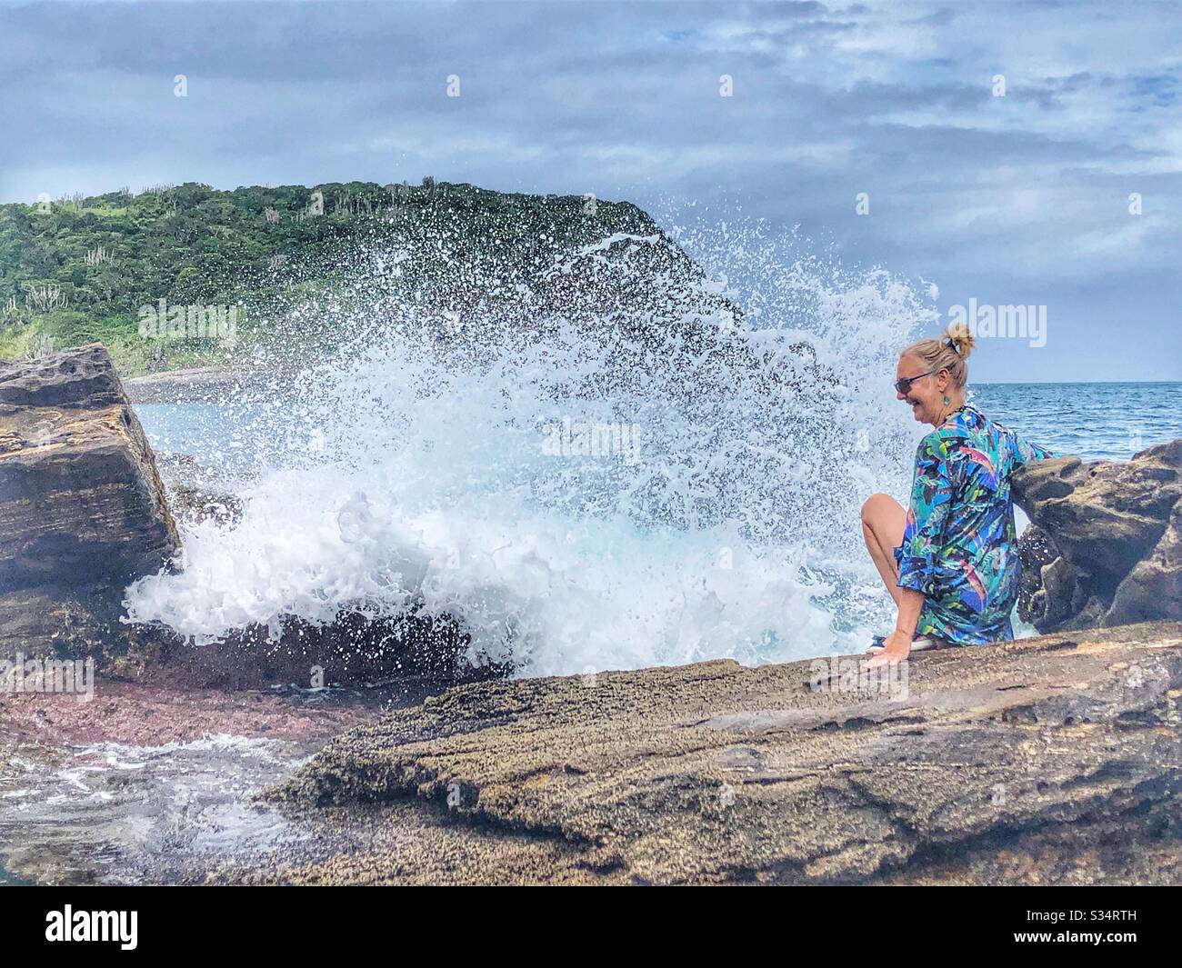 Woman sitting on a rock gets splashed by the spray of a wave. - Smartphone Captured Stock Image