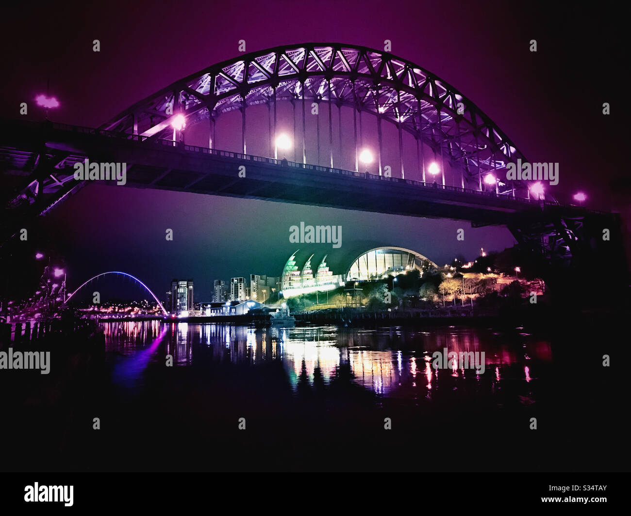 Architecture of Newcastle Quayside lit up by night. Newcastle upon Tyne, North East England. The Tyne Bridge with Sage Gateshead and Millennium Bridge illuminated with colourful lights - Smartphone Captured Stock Image