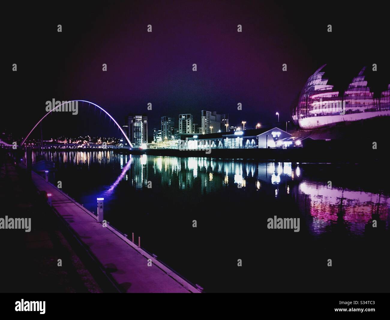 Architecture of Newcastle Quayside lit up by night. Newcastle upon Tyne, North East England. Colourful city lights reflecting in the River Tyne with Sage Gateshead and Millennium Bridge - Smartphone Captured Stock Image