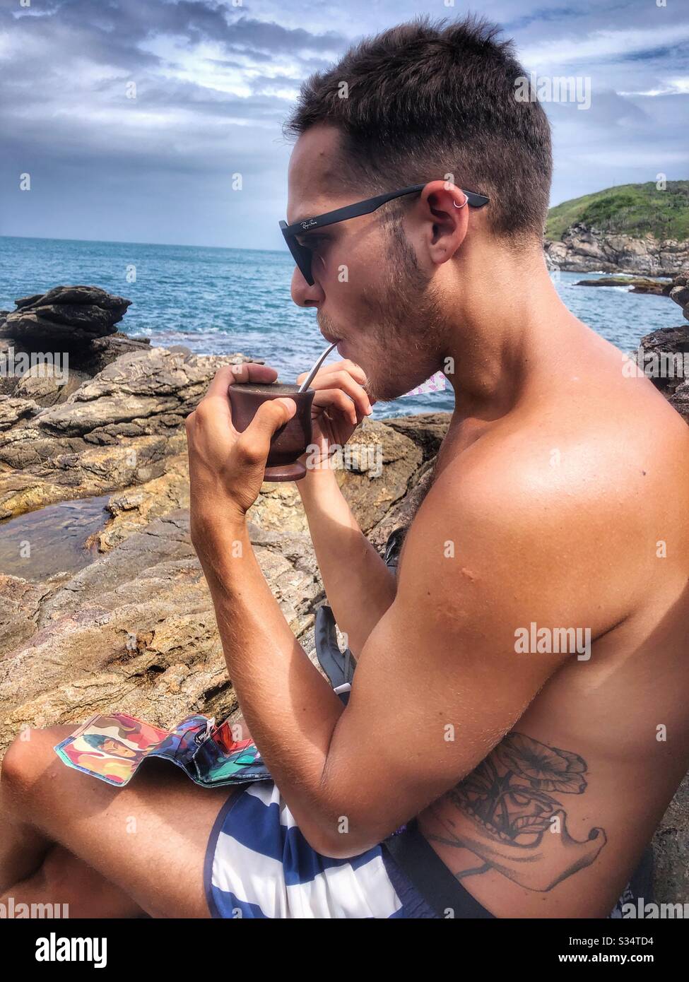 A young man relaxing by the sea drinking Maté on the rocky shoreline. - Smartphone Captured Stock Image