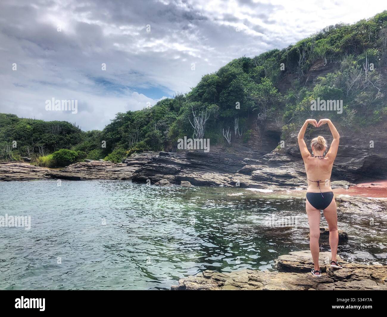 Woman makes the shape of a heart with her arms while standing on the rocks by the sea. Stock Photo