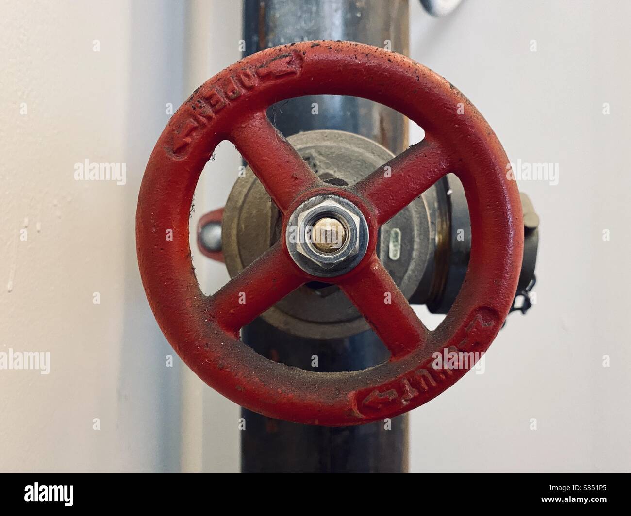 Red, metal valve wheel with open and shut arrows, on black pipe next to white walls - Smartphone Captured Stock Image