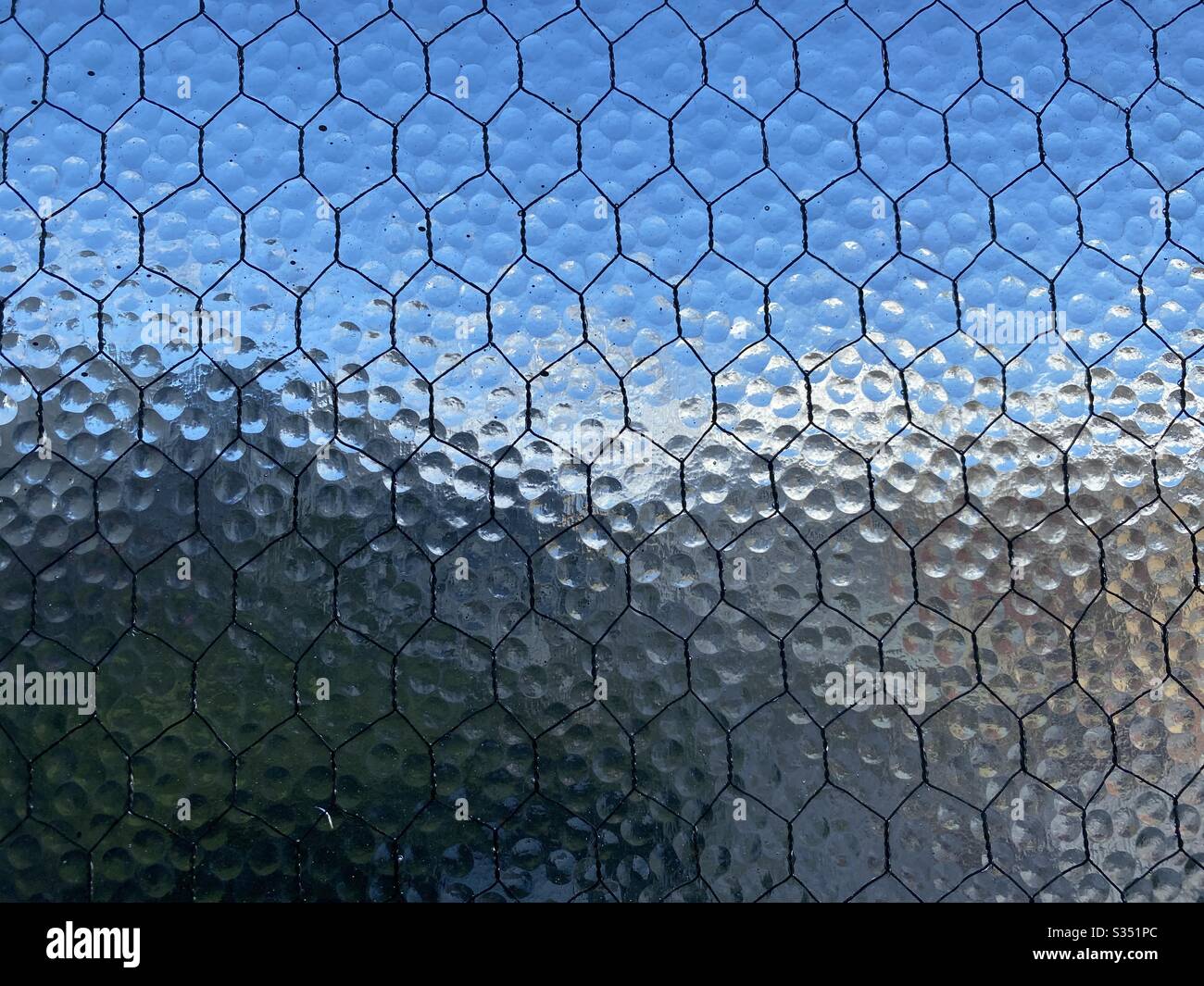 Stippled, opaque safety glass with wire mesh running through it, Indistinct blue sky and city barely visible through it, creating abstract background texture. - Smartphone Captured Stock Image