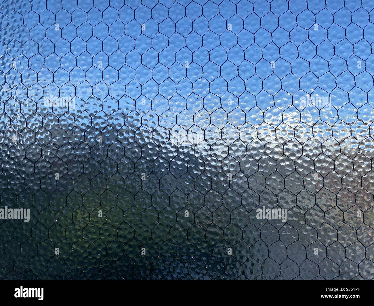 Wide view stippled, opaque safety glass with wire mesh running through it, Indistinct blue sky and city barely visible through it, creating abstract background texture. - Smartphone Captured Stock Image