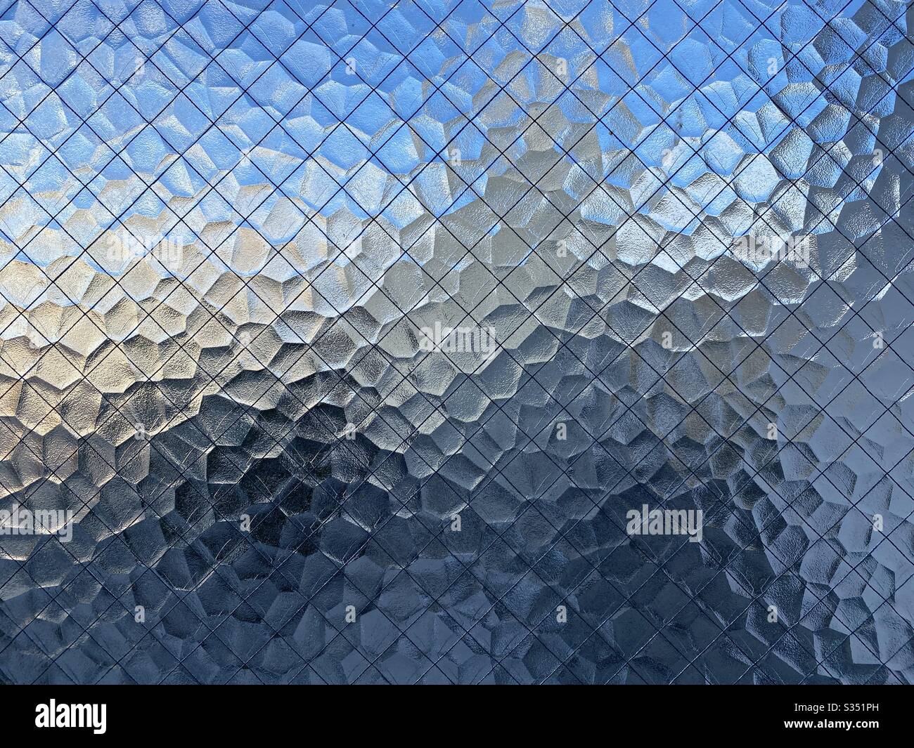 Textured, opaque safety glass with wire mesh running through it, Indistinct blue sky and city barely visible through it, creating abstract background texture. - Smartphone Captured Stock Image