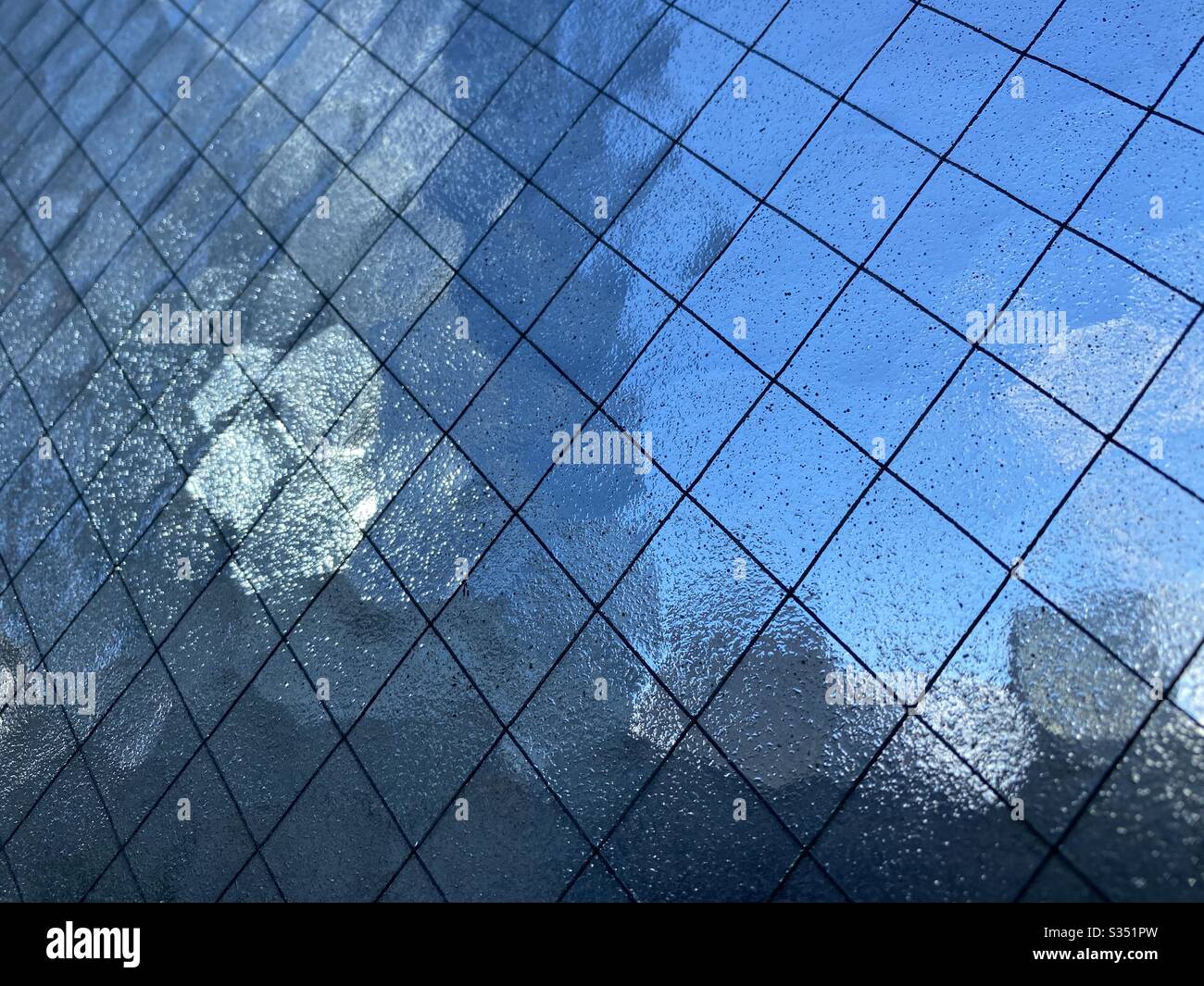 Close, angled view of opaque safety glass with wire mesh running through it, Indistinct blue sky and light reflecting off nearby building visible through it, creating abstract background texture. - Smartphone Captured Stock Image