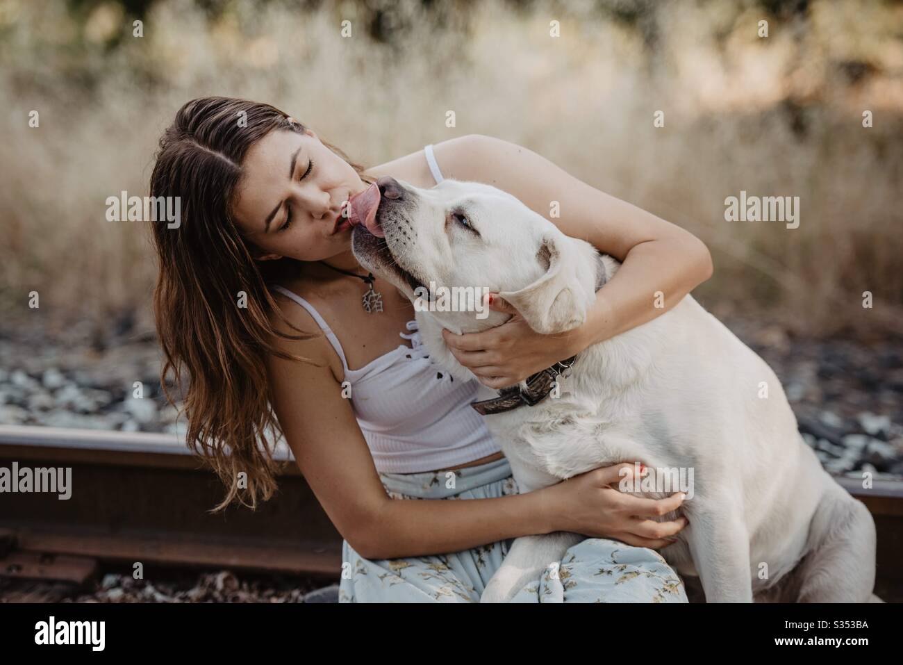 Girl getting kisses from white lab and wearing a white tank top sitting on train tracks. Stock Photo