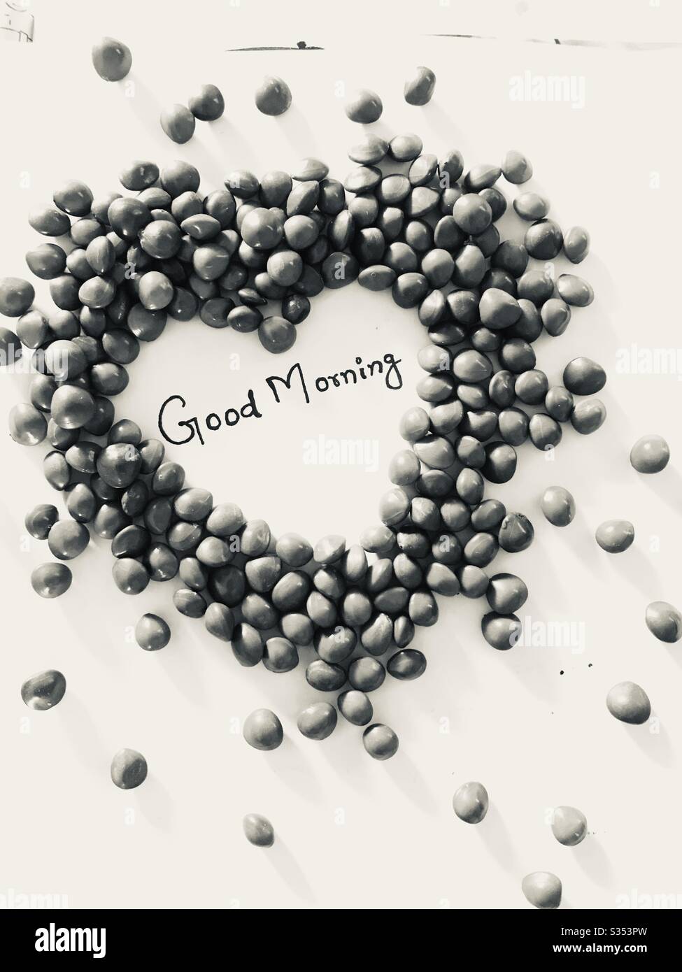 Adenanthera red beads in shape of heart with words Good Morning hand written, with white surface background. Morning message for friends,Manjadikuru in malayalam,black & white mode Stock Photo