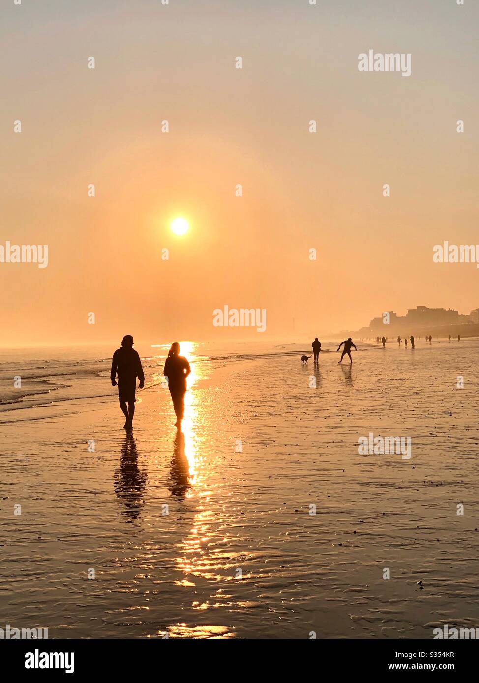 A couple walk silhouetted against the golden setting sun on a beach at low tide with other walkers in the background Stock Photo