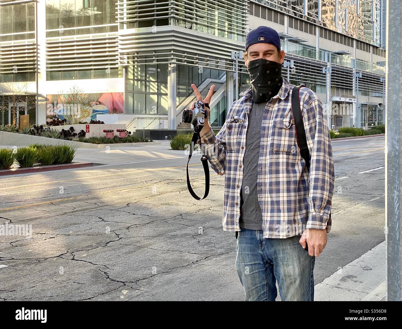Photographer wearing bandana to cover face, gives victory sign between taking pictures on streets in Downtown Los Angeles during coronavirus, Covid-19 pandemic. Identifying logos and signage removed - Smartphone Captured Stock Image