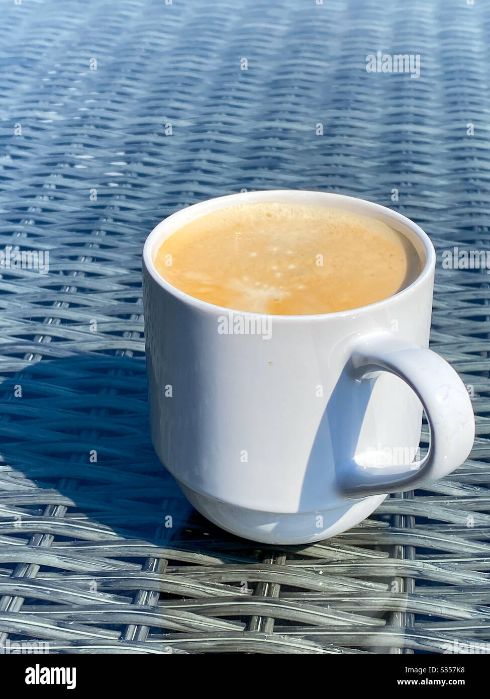 White mug of hot frothy coffee on the glass table top of garden furniture Stock Photo