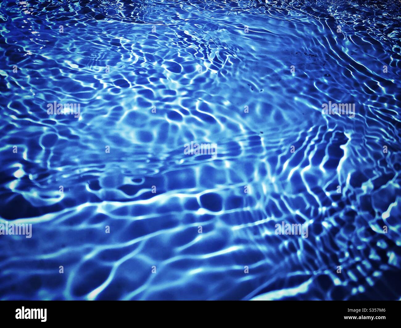 A close up view on the surface of water in a blue swimming pool. Light reflects off gentle ripples creating a colourful summer background pattern. Fresh, clean and vibrant summery vibes. Stock Photo