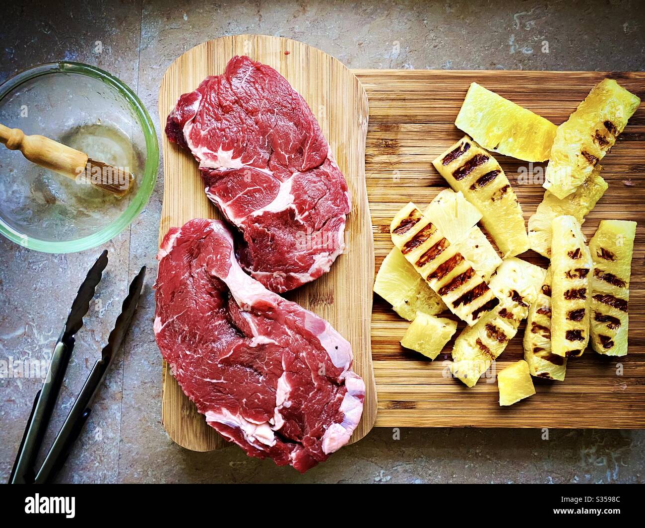 Preparing dinner in the kitchen. Fresh food preparation. Ingredients for homemade pineapple salsa. Chargrilled slices on a wooden chopping board with two ribeye steaks and honey marinade - Smartphone Captured Stock Image