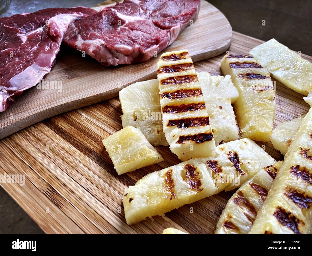 Preparing dinner in the kitchen. Fresh food preparation. Ingredients for homemade pineapple salsa. Chargrilled slices on a wooden chopping board with two ribeye steaks - Smartphone Captured Stock Image