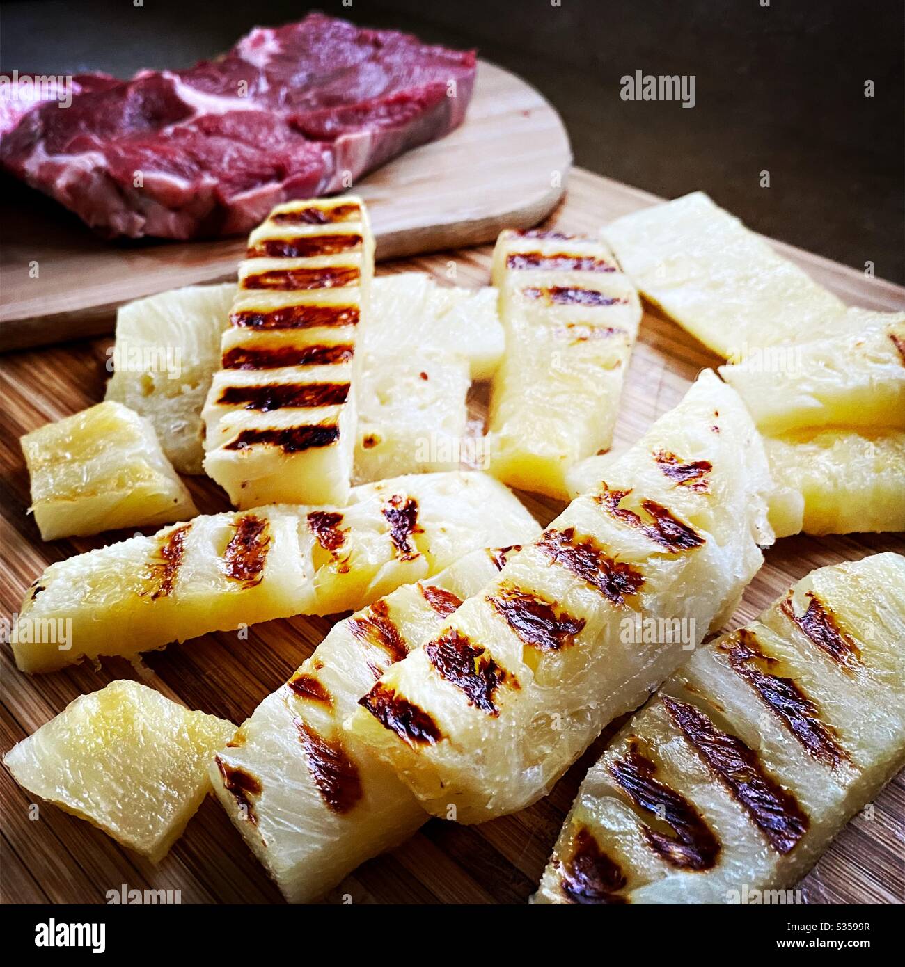 Preparing dinner in the kitchen. Fresh food preparation. Ingredients for homemade pineapple salsa. Chargrilled slices on a wooden chopping board with two ribeye steaks - Smartphone Captured Stock Image