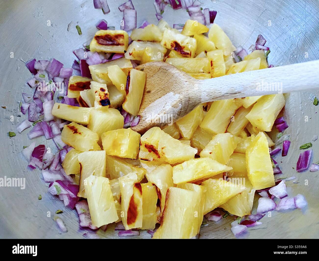 Preparing pineapple dish in the kitchen. Fresh and ripe fruit preparation. Ingredients for homemade pineapple salsa. Chargrilled chunks with red onion, lime juice and coriander in a steel mixing bowl. - Smartphone Captured Stock Image
